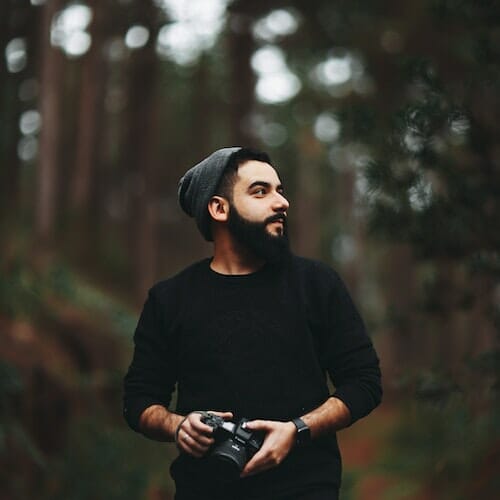 A bearded man holding a camera in the woods.